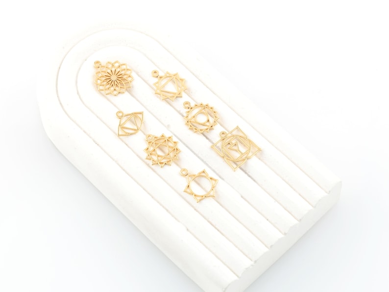 May include: Seven gold-colored metal charms with different geometric designs, including a flower, a triangle, and a square. The charms are arranged on a white surface.