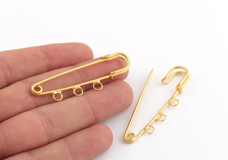 May include: Two gold-toned safety pin closures with four jump rings on each. The safety pins are open and lying on a hand.