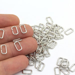 May include: A pile of silver-toned metal D-rings, also known as jump rings, are scattered on a white surface. A hand is holding a few of the rings.