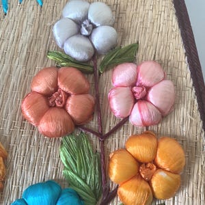 Puede incluir: Primer plano de un dise&ntilde;o floral decorativo. La obra presenta flores en tonos naranja, rosa, amarillo y blanco, con hojas verdes, todo sobre un fondo de paja tejida. Las flores est&aacute;n hechas de un material sedoso.