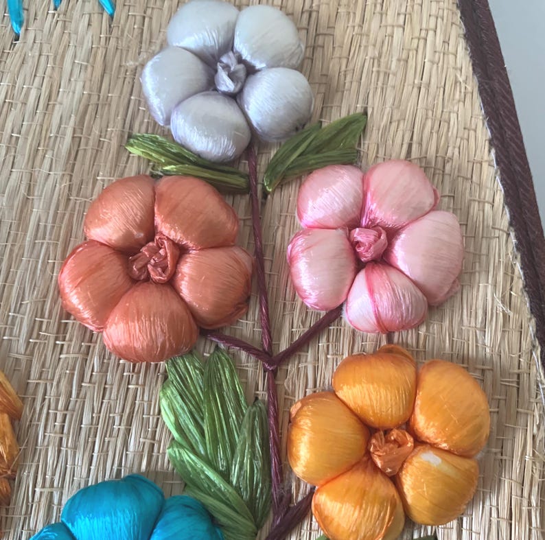 Puede incluir: Primer plano de un dise&ntilde;o floral decorativo. La obra presenta flores en tonos naranja, rosa, amarillo y blanco, con hojas verdes, todo sobre un fondo de paja tejida. Las flores est&aacute;n hechas de un material sedoso.