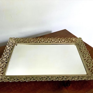 May include: A rectangular gold-toned vanity tray with a mirrored surface. The tray features an ornate, openwork design around the edges. The tray is displayed on a wooden surface.