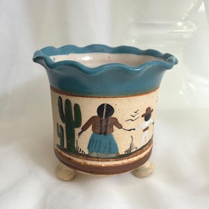 May include: A ceramic planter with a scalloped blue rim and three small feet. The beige body features a hand-painted scene of a green cactus and two figures, one in a brown top and blue skirt, the other in a white outfit and brown hat.