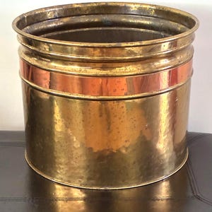 May include: A cylindrical brass planter featuring a polished copper band encircling the upper section. The metal surface displays a subtle hammered texture with visible signs of natural patina and aging throughout the exterior finish.