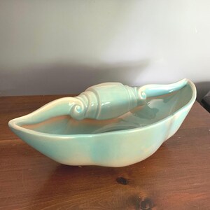 May include: A ceramic bowl with a glossy light blue and cream glaze, featuring an elongated, leaf-like shape and a central handle that arches over the middle, resembling a stylized shell or boat with smooth, curved edges and a reflective finish.