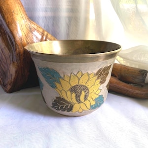 May include: A decorative bowl with a brass rim and a painted floral design. The bowl features a large yellow sunflower with brown and teal leaves against a cream background. The interior and rim are a shiny brass colour.
