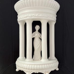 May include: A white matte ceramic decorative piece featuring a central standing figure enclosed within a structure of four fluted columns. The base and top are adorned with scalloped edges and embossed detailing, creating a temple-like appearance.