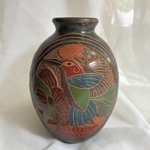 May include: A dark brown ceramic vase featuring a hand-painted relief design of a hummingbird in flight surrounded by large flowers and foliage. The bird is depicted with red, blue, and green accents, while the background texture is rough and matte.