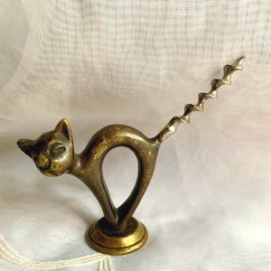 May include: A vintage brass cat corkscrew. The cat is arched with a spiral tail, and its head is turned. The corkscrew is mounted on a round base. The metal has a warm, antique finish, and the piece is likely made of brass.