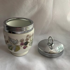 K&ouml;nnte beinhalten: Eine wei&szlig;e Keramik-Eierbecher mit einem silbernen Deckel und einem Blumendesign mit Brombeeren. Der Deckel hat einen silbernen Griff.