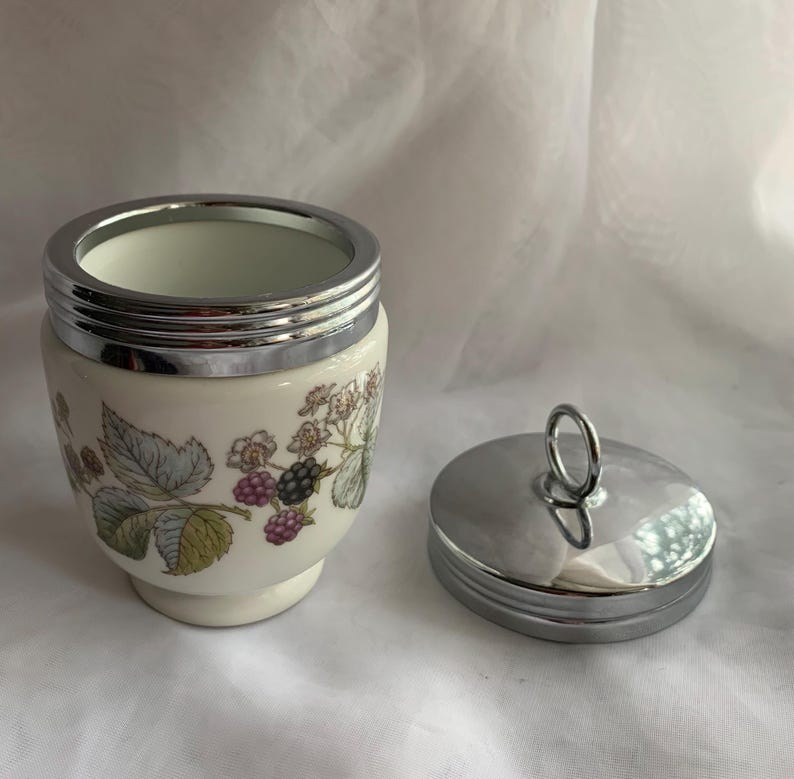 K&ouml;nnte beinhalten: Eine wei&szlig;e Keramik-Eierbecher mit einem silbernen Deckel und einem Blumendesign mit Brombeeren. Der Deckel hat einen silbernen Griff.
