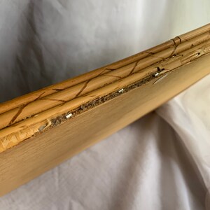 May include: Close-up of a light brown wooden picture frame with a bamboo-style border. The frame shows signs of wear, with some areas of the wood chipped and staples visible. The frame is angled, revealing its construction and texture.
