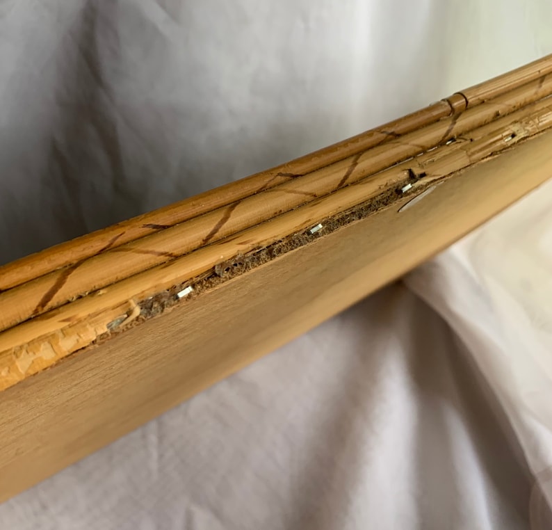 May include: Close-up of a light brown wooden picture frame with a bamboo-style border. The frame shows signs of wear, with some areas of the wood chipped and staples visible. The frame is angled, revealing its construction and texture.