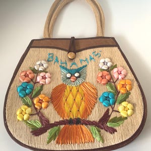 Puede incluir: Un bolso de mano de paja con ribetes y asas marrones. El bolso presenta un dise&ntilde;o de b&uacute;ho bordado con flores coloridas y la palabra "Bahamas". El b&uacute;ho tiene detalles azules, naranjas y amarillos.