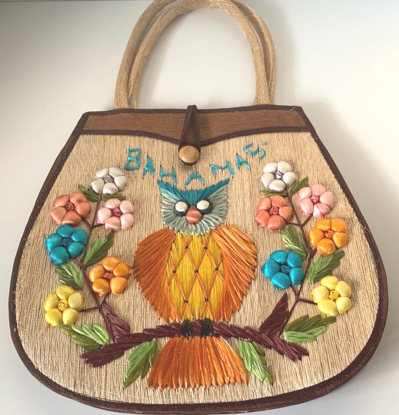 Puede incluir: Un bolso de mano de paja con ribetes y asas marrones. El bolso presenta un dise&ntilde;o de b&uacute;ho bordado con flores coloridas y la palabra "Bahamas". El b&uacute;ho tiene detalles azules, naranjas y amarillos.