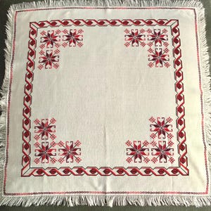 May include: White square tablecloth with red and black embroidered geometric patterns and fringe trim. The design includes corner starburst motifs and a border of interlocking shapes. A traditional textile.