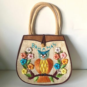 Puede incluir: Bolso de mano vintage con textura de paja tejida, con un dise&ntilde;o de b&uacute;ho bordado. El bolso tiene un marco de madera marr&oacute;n, un cierre de bot&oacute;n y asas tejidas. La palabra "Bahamas" est&aacute; bordada sobre el b&uacute;ho, rodeada de flores coloridas.