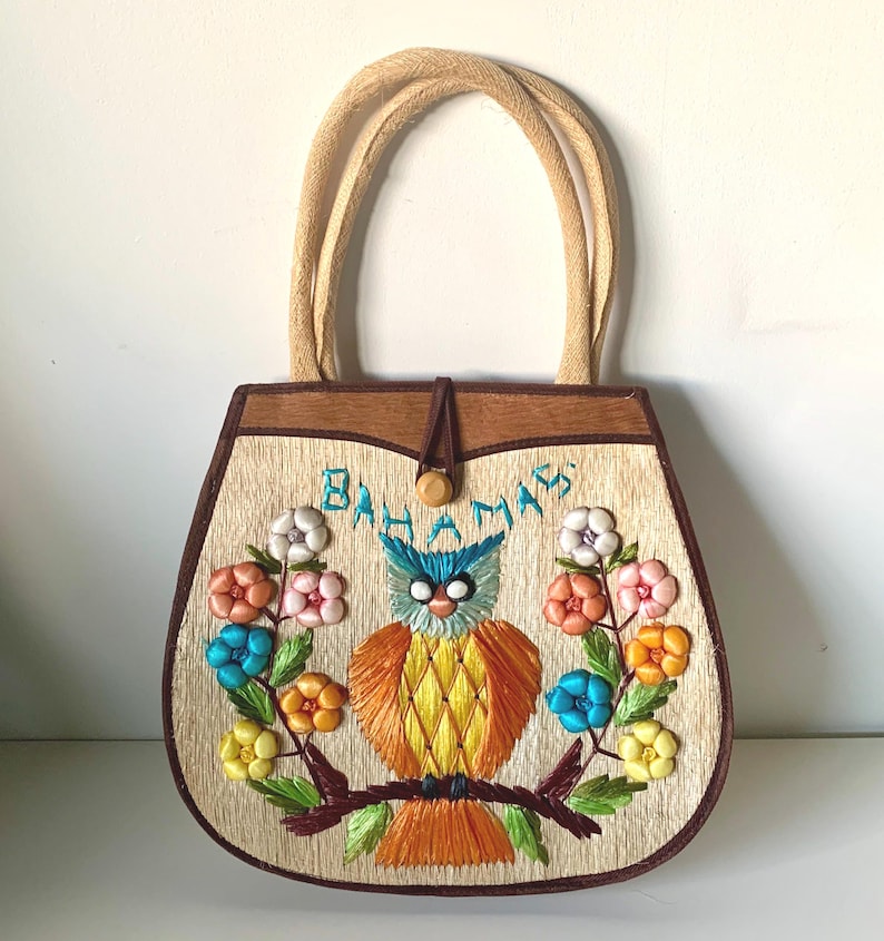 Puede incluir: Bolso de mano vintage con textura de paja tejida, con un dise&ntilde;o de b&uacute;ho bordado. El bolso tiene un marco de madera marr&oacute;n, un cierre de bot&oacute;n y asas tejidas. La palabra "Bahamas" est&aacute; bordada sobre el b&uacute;ho, rodeada de flores coloridas.