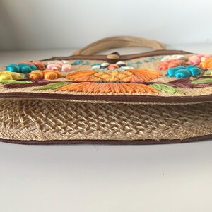 Puede incluir: Un bolso de mano de paja tejida con asa y ribete marr&oacute;n. El bolso presenta flores bordadas de colores en tonos naranja, amarillo y azul. El bolso tiene un cierre de bot&oacute;n y un cuerpo de paja tejida.