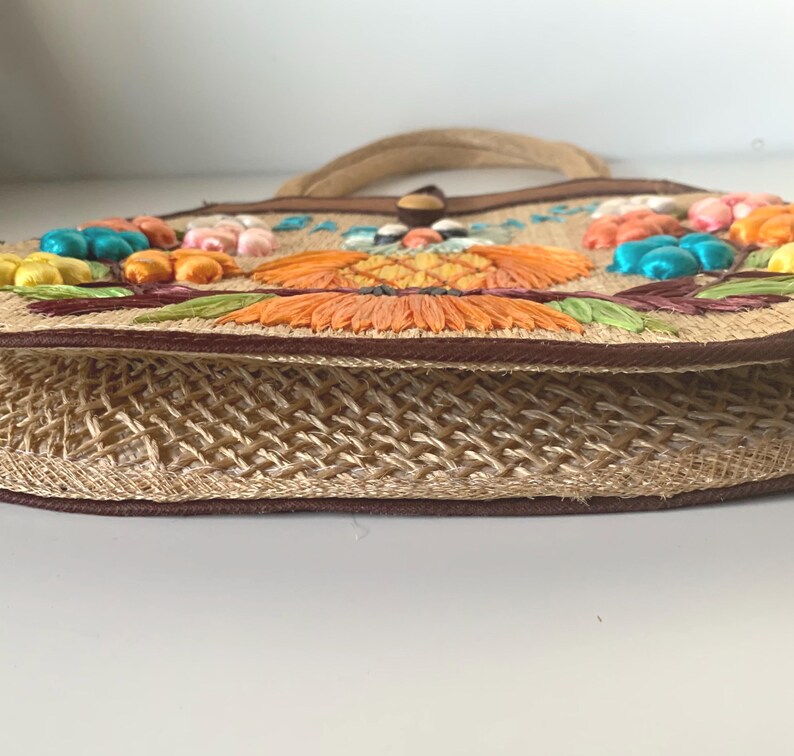 Puede incluir: Un bolso de mano de paja tejida con asa y ribete marr&oacute;n. El bolso presenta flores bordadas de colores en tonos naranja, amarillo y azul. El bolso tiene un cierre de bot&oacute;n y un cuerpo de paja tejida.