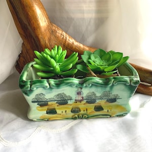 May include: A rectangular ceramic planter with a green trim and a landscape design. The planter holds two artificial green succulents. The planter's design features a woman in a red and white dress. The planter is on a white surface.