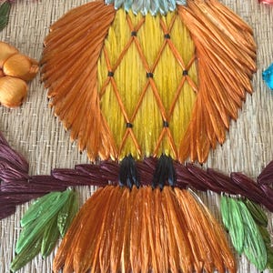 Puede incluir: Una colorida obra de arte de un p&aacute;jaro hecha a mano. El p&aacute;jaro presenta tonos naranja, amarillo y azul, con una textura tejida detallada. Est&aacute; posado en una rama marr&oacute;n con hojas verdes, sobre un fondo tejido natural.