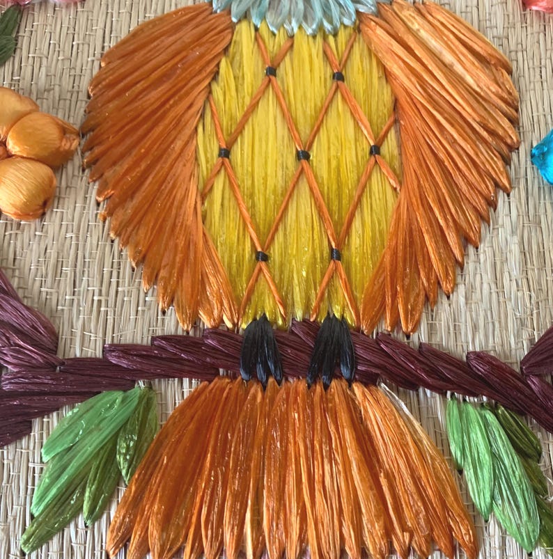 Puede incluir: Una colorida obra de arte de un p&aacute;jaro hecha a mano. El p&aacute;jaro presenta tonos naranja, amarillo y azul, con una textura tejida detallada. Est&aacute; posado en una rama marr&oacute;n con hojas verdes, sobre un fondo tejido natural.