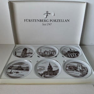 May include: A set of six white porcelain coasters featuring black and white illustrations of buildings. The coasters are presented in a white box with the text "Fürstenberg Porzellan Seit 1747".