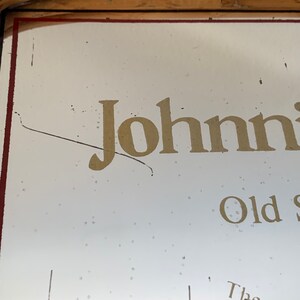 May include: A vintage sign with the name "Johnnie" in gold lettering. The sign has a white background with a red border and a wooden frame. The words "Old S" are also visible.