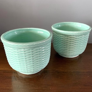 May include: Two round ceramic planters in a light mint green colour. Each planter features a textured exterior surface that mimics the appearance of a woven basket pattern, with a smooth interior finish and a simple, circular rim design.