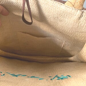 Puede incluir: Vista interior de un bolso tote tejido beige. El bolso tiene un asa marr&oacute;n y un peque&ntilde;o bolsillo. El interior est&aacute; forrado con una tela beige. La costura turquesa decora la base del bolso.