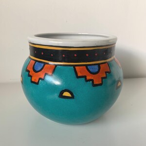 Peut inclure: Un pot en céramique arrondi avec une base turquoise vive ornée de formes géométriques peintes à la main en orange et bleu. Le bord supérieur présente une large bande noire avec des points orange, et la partie inférieure inclut des demi-cercles jaunes.