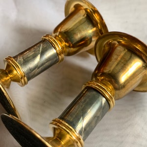 Op de afbeelding: Een paar gouden en zilveren kandelaars met een gedraaid ontwerp op de basis. De kandelaars zijn gemaakt van metaal en hebben een gepolijste afwerking.