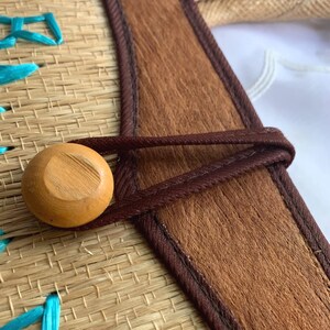 Puede incluir: Primer plano de un bolso de paja tejido con una correa de cuero marr&oacute;n y un cierre de bot&oacute;n de madera. El bolso presenta costuras turquesas y un aspecto natural y texturizado. La correa de cuero tiene una textura marr&oacute;n similar a la piel.