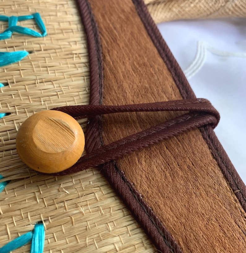 Puede incluir: Primer plano de un bolso de paja tejido con una correa de cuero marr&oacute;n y un cierre de bot&oacute;n de madera. El bolso presenta costuras turquesas y un aspecto natural y texturizado. La correa de cuero tiene una textura marr&oacute;n similar a la piel.
