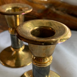 Op de afbeelding: Twee gouden kandelaars met zwarte marmeren voetstukken. De kandelaars hebben een eenvoudig ontwerp met een ronde bovenkant en een cilindrische voet.