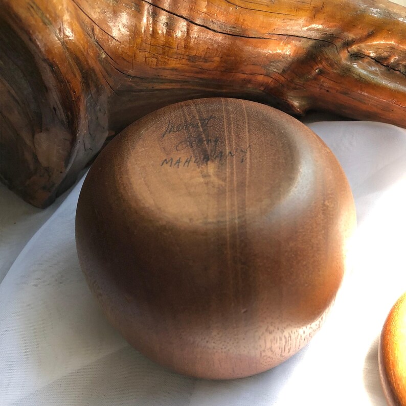 以下が含まれることがあります： 手作りの丸いマホガニーの木製ボウル。ボウルは滑らかで磨かれた表面と自然な木目模様を持っています。ボウルの底には「Mary C. Mahogany」の文字が刻まれています。