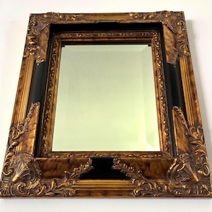 May include: A rectangular wall mirror featuring a thick, ornate frame with a combination of metallic gold and matte black finishes. The frame displays intricate raised scrollwork and leaf-like carvings at the corners and edges, surrounding a clear, reflective glass pane with beveled edges.
