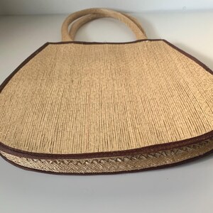 Puede incluir: Un bolso tote de paja tejida con ribete y asas marrones. El bolso tiene forma trapezoidal y un borde cosido decorativo. Las asas est&aacute;n hechas de un material de cuerda trenzada.