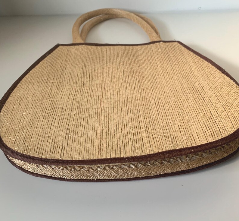 Puede incluir: Un bolso tote de paja tejida con ribete y asas marrones. El bolso tiene forma trapezoidal y un borde cosido decorativo. Las asas est&aacute;n hechas de un material de cuerda trenzada.