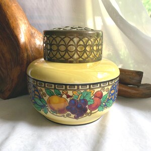 May include: A yellow ceramic jar with a brass floral frog lid. The jar features a colorful fruit and leaf design around the base. The lid has a decorative openwork pattern. The jar is likely for floral arrangements.