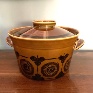 May include: A ceramic cooking pot with a lid. The pot is a warm, golden-brown colour with a darker brown lid and handles. The pot is decorated with a repeating pattern of stylised floral designs in a dark brown colour.