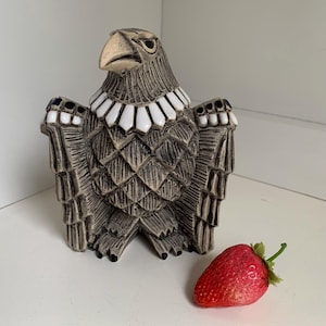 May include: A ceramic eagle sculpture with a white and black collar, standing on a white surface with a red strawberry in front of it.