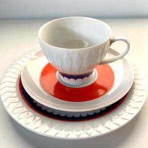 May include: A white teacup with a textured design sits on a saucer with a bright orange centre and a decorative blue and purple border. The set rests on a larger white plate with a repeating leaf design, creating a layered effect.