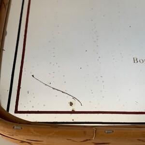 May include: A rectangular mirror with a light brown wooden frame. The mirror's surface is white with a few dark spots and a long, thin, dark hair-like object. The frame has a dark red and black border.