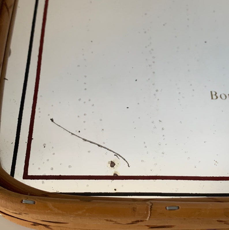 May include: A rectangular mirror with a light brown wooden frame. The mirror's surface is white with a few dark spots and a long, thin, dark hair-like object. The frame has a dark red and black border.