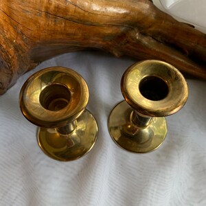 Op de afbeelding: Twee gouden metalen kaarsenstandaards met een eenvoudig ontwerp. De kaarsenstandaards staan op een wit oppervlak.