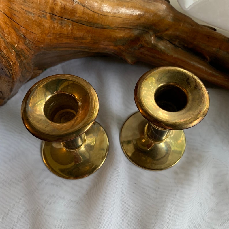 Op de afbeelding: Twee gouden metalen kaarsenstandaards met een eenvoudig ontwerp. De kaarsenstandaards staan op een wit oppervlak.