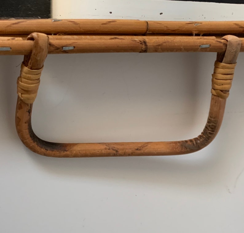 May include: Close-up of a bamboo mirror with a curved handle. The handle is made of bent bamboo and wrapped with woven material at the joints. The mirror frame is made of bamboo and secured with small metal staples.
