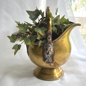 May include: A vintage brass coal scuttle with a handle, filled with faux green ivy. The scuttle has a decorative face on the front and sits on a round base. The brass has a warm, aged patina.